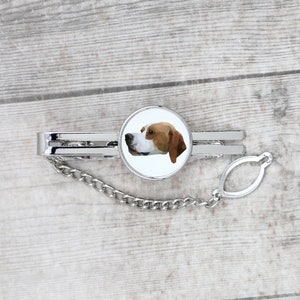 May include: A silver tie clip with a white circular design featuring a brown and white dog head. The tie clip has a silver chain attached.