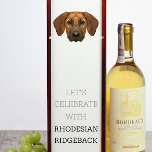 May include: A wooden wine box with a white rope handle. The box has a white label with the text "Let's Celebrate with Rhodesian Ridgeback" and a geometric illustration of a Rhodesian Ridgeback dog. A bottle of white wine is next to the box.