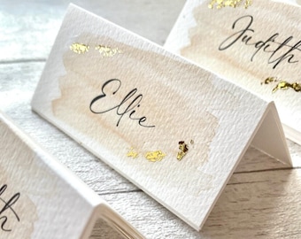 Könnte beinhalten: Gefaltete weiße Tischkarten mit aquarellartigen beigen Akzenten und Goldfoliendetails. Der Name "Elfie" ist in eleganter Kalligraphie geschrieben. Weitere Karten mit Namen sind sichtbar, was auf ein Hochzeits- oder Eventthema hindeutet.