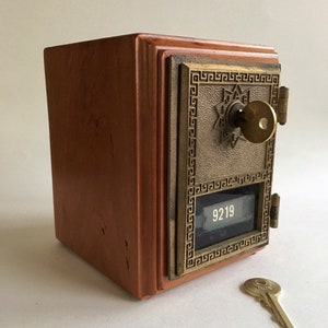 May include: A vintage wooden mailbox with a brass door and a key in the lock. The door has a decorative design and a small window with the number 9219 visible. A brass key is lying on the table next to the mailbox.