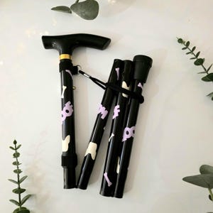 May include: A black folding walking stick with a black handle and a gold accent. The stick has a pattern of white and lavender bone shapes. The stick is folded and secured with a black strap.