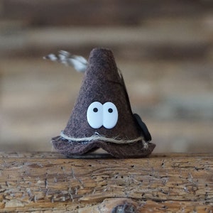 Puede incluir: Un sombrero de fieltro marr&oacute;n en forma de cono con ojos blancos y una pluma que sobresale de la parte superior. El sombrero est&aacute; sobre una superficie de madera.