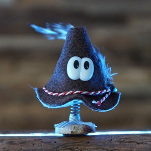 Puede incluir: Un sombrero de fieltro marr&oacute;n con ojos blancos y un borde rojo y blanco. El sombrero est&aacute; en un resorte y est&aacute; sentado sobre una superficie de madera.