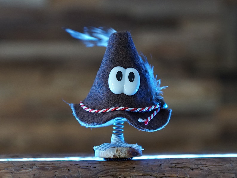 Puede incluir: Un sombrero de fieltro marr&oacute;n con ojos blancos y un borde rojo y blanco. El sombrero est&aacute; en un resorte y est&aacute; sentado sobre una superficie de madera.