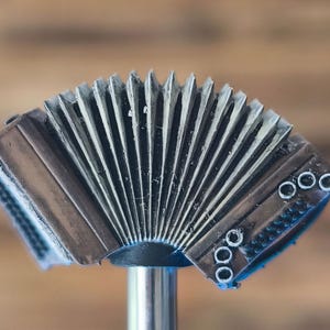 Op de afbeelding: Een miniatuur accordeon met bruine en zilveren tinten. De balg is zilverkleurig en de behuizing is bruin met zilveren details. Het is gemonteerd op een zilveren standaard. De achtergrond is een vage bruine kleur.