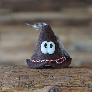 Puede incluir: Un sombrero de fieltro marr&oacute;n con una pluma blanca y un ribete rojo y blanco. El sombrero tiene dos grandes ojos saltones blancos.