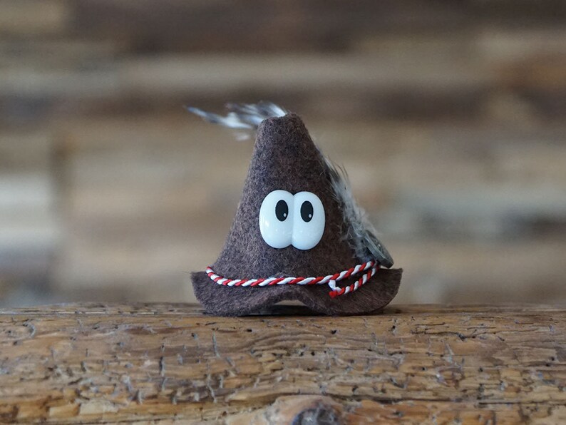 Puede incluir: Un sombrero de fieltro marr&oacute;n con una pluma blanca y un ribete rojo y blanco. El sombrero tiene dos grandes ojos saltones blancos.