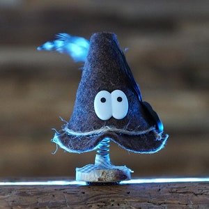 Puede incluir: Un gnomo de fieltro marr&oacute;n con ojos saltones blancos y una pluma negra en la parte superior de su gorro. El gnomo est&aacute; sentado en un resorte y est&aacute; sobre una superficie de madera.