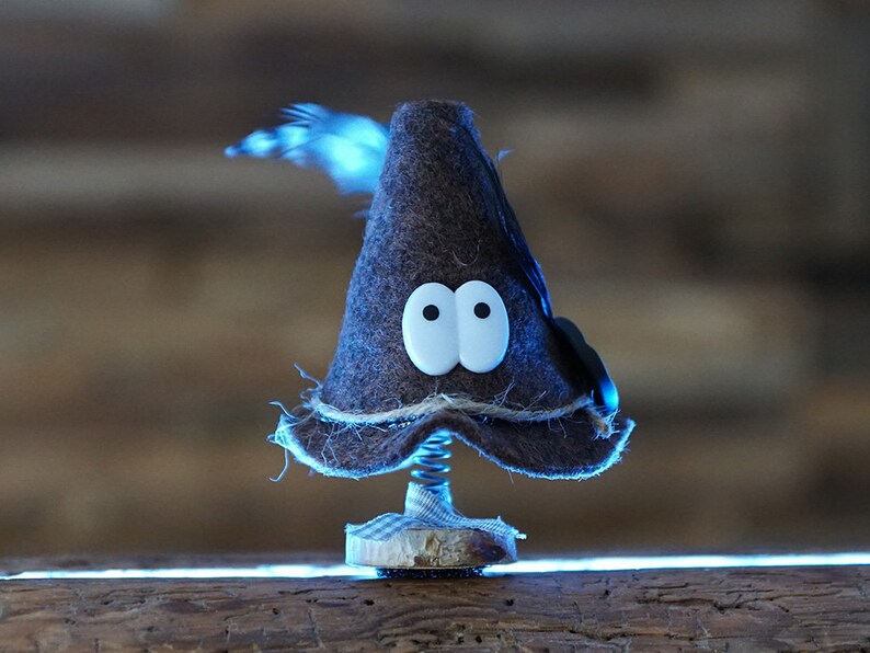 Puede incluir: Un gnomo de fieltro marr&oacute;n con ojos saltones blancos y una pluma negra en la parte superior de su gorro. El gnomo est&aacute; sentado en un resorte y est&aacute; sobre una superficie de madera.