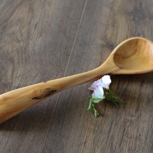 Può includere: Un cucchiaio di legno fatto a mano con un lungo manico e una ciotola arrotondata. Il cucchiaio è fatto di legno chiaro e poggia su una superficie di legno. Un piccolo rametto di fiori rosa e bianchi è accanto al cucchiaio.