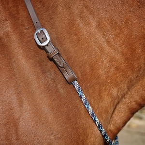 May include: A brown leather strap with a silver metal buckle and a keeper loop, connected to a braided rope in shades of blue, white, and black. The leather component features a small embossed logo and is positioned against a brown animal coat.