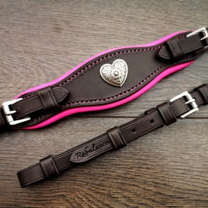 May include: Two-piece equestrian tack set. The larger piece is dark brown leather with a bright pink underside and a silver heart-shaped embellishment. The smaller piece is dark brown leather with the word "Rebalance" embossed on it. Both pieces have silver buckles.