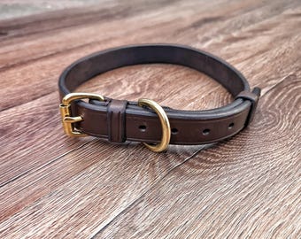 May include: A brown leather dog collar with a gold-colored buckle and D-ring. The collar is circular and has a classic design. The collar is approximately 1 inch wide.