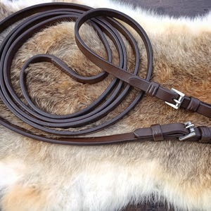 Puede incluir: Una rienda de caballo de cuero marrón enrollada, con hebillas plateadas. La rienda está sobre una superficie texturizada marrón y beige, probablemente piel. El cuero parece liso y bien cuidado.