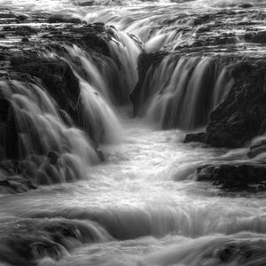 Op de afbeelding: Zwart-wit foto van een waterval. Het water stroomt over donkere rotsformaties, waardoor een dynamische en gestructureerde scène ontstaat. De afbeelding legt de beweging en kracht van het water vast.