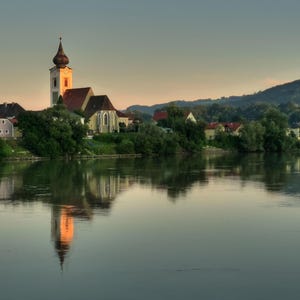 Op de afbeelding: Schilderachtig landschap met een kerk met een hoge torenspits die weerspiegeld wordt in kalm water. De kerk heeft een rood dak en is omgeven door groene bomen en andere gebouwen. De lucht vertoont een zachte kleurovergang.