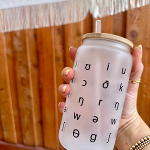 May include: A frosted white reusable water bottle with a bamboo lid and a clear straw. The bottle has black letters printed on it, including the letters "i", "u", "o", "k", "n", "r", "w", "a", "e", "g", and "j".