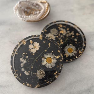 Puede incluir: Dos posavasos de resina negra con destellos dorados y flores blancas prensadas. Los posavasos son redondos y tienen un acabado brillante.