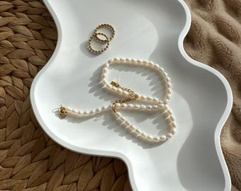 May include: A white, wavy-edged tray holds a pearl necklace and two gold rings. The necklace has a gold clasp. The rings are gold and have a textured design. The tray sits on a woven mat and a soft, tan blanket.