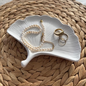May include: A white, fan-shaped ceramic dish holds a pearl necklace and several gold rings. The dish sits on a woven, light brown surface. The necklace is made of small, round pearls with a gold clasp.