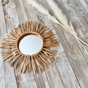 May include: A round mirror with a sunburst design made of woven brown paper strips. The mirror is hanging on a whitewashed wood background.
