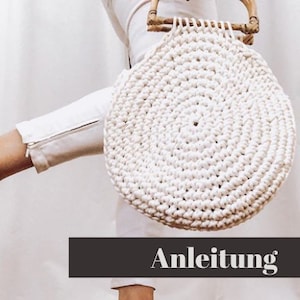 May include: A white crocheted round bag with a wooden handle. The bag is made with a circular pattern and has a natural, rustic look. The text "Anleitung" is visible in the image.