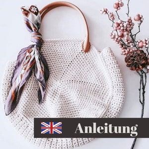 May include: A white crocheted tote bag with a brown leather handle and a purple and brown patterned scarf draped over the top. The bag has a crocheted texture and the text "Anleitung" is printed on a black bar below the bag.