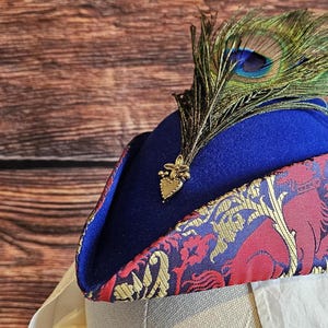 Può includere: Un cappello di feltro blu con una fascia a motivi rossi e dorati e un accento di piuma di pavone. Il cappello ha un ornamento a forma di cuore dorato.