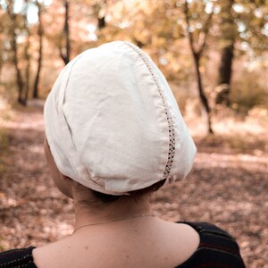 May include: A white linen cap with a decorative stitch detail. The cap is worn by a person standing in a forest setting.