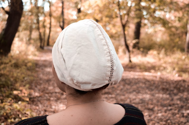 May include: A white linen cap with a decorative stitch detail. The cap is worn by a person standing in a forest setting.