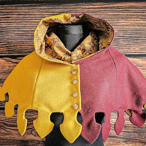 May include: A two-toned hooded capelet featuring a mustard yellow left side and a deep burgundy right side. The garment has a vertical row of four metallic buttons down the center and a jagged, pointed hemline. The interior lining has a gold pattern.