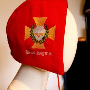以下が含まれることがあります： 前面に刺繍されたドクロと交差した骨の模様が施された赤い布の帽子。帽子には、後ろに青と赤の紐が付いています。ドクロと交差した骨の下には「Deus Sigmar」という文字が刺繍されています。
