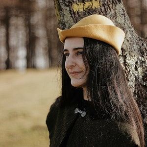 May include: A woman with long dark hair wearing a yellow felt hat and a black jacket with a silver brooch. She is standing in front of a tree with white markings.