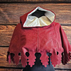 Peut inclure: Une cape à capuche rouge foncé avec une doublure intérieure de couleur claire et un ourlet festonné présentant des découpes en forme de feuilles sur le bord inférieur. Le vêtement est présenté sur un mannequin, mettant en valeur la capuche.