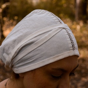 May include: A white linen cap with a folded design and a delicate lace trim. The cap is worn on a person's head.