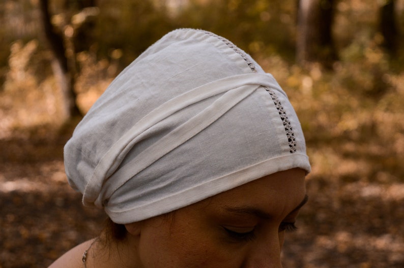 May include: A white linen cap with a folded design and a delicate lace trim. The cap is worn on a person's head.