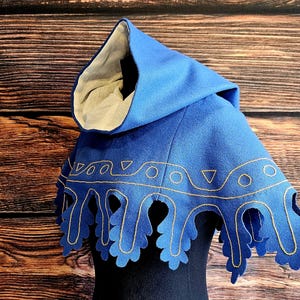 May include: A blue hooded cloak with gold trim. The cloak features a pointed hood and a scalloped edge with gold embroidery. The hood's interior is a light beige colour. The cloak is a medieval style.