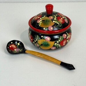 May include: A black wooden lidded jar and a matching spoon, both decorated with hand-painted red, yellow, and green floral motifs. The jar features a rounded body, a red rim, and a red knob on the lid, while the spoon has a long, light-colored handle.