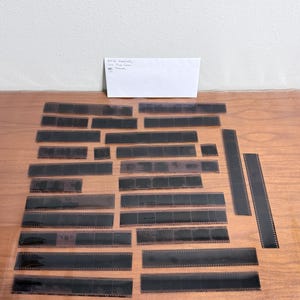 May include: A collection of black film negatives of varying lengths, arranged on a wooden surface. A white paper with handwritten text is in the background. The negatives are likely for photography or film processing.