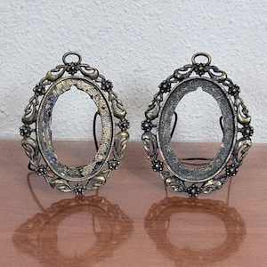 May include: Two oval-shaped metal picture frames with an antique bronze finish. Each frame features an ornate, decorative border with small floral accents and a top loop. The frames are designed to stand upright with a thin metal support.