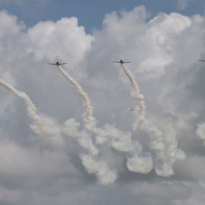 Puede incluir: Cinco pequeños aviones vuelan en formación, dejando estelas de humo blanco en un cielo azul con nubes blancas.