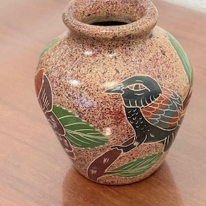 Könnte beinhalten: Eine kleine, runde Keramikvase mit einer gesprenkelten, braun-beigen, strukturierten Oberfläche. Das Gefäß ist mit handgemalten Illustrationen von schwarzen Vögeln verziert, die auf braunen Zweigen mit grünen Blättern sitzen.