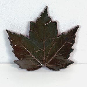 May include: A ceramic dish shaped like a maple leaf with a glossy, dark brown and forest green glaze. The surface features detailed, embossed veins that mimic the natural structure of a leaf, with a smooth, reflective finish across the entire piece.