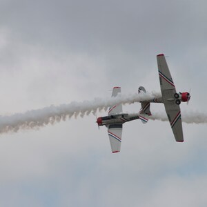 Puede incluir: Dos aviones blancos con detalles rojos y azules vuelan en formación, dejando un rastro de humo blanco detrás de ellos.