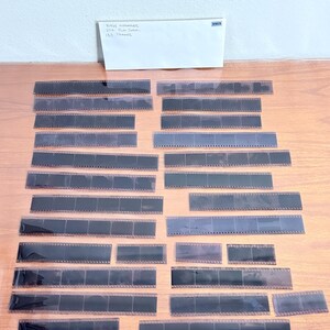 May include: A collection of black and translucent film negatives arranged on a wooden surface. A white envelope with handwritten text is in the background. The text on the envelope reads "BMW Netherlands V2A Film Series 153 Frames".