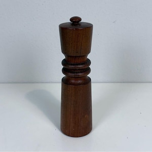 Może przedstawiać: Ciemnobrązowy drewniany młynek do pieprzu. Ma cylindryczną podstawę, żebrowaną środkową część i stożkową górę z małym pokrętłem. Młynek do pieprzu jest wykonany z ciemnego drewna i ma gładkie wykończenie. To przybory kuchenne.