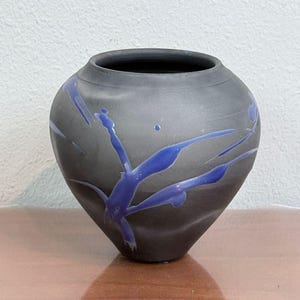 May include: A matte black ceramic vase with a rounded body and a narrow opening. The surface features a striking, abstract blue glaze pattern that resembles fluid, organic splashes or streaks extending across the dark, textured exterior.