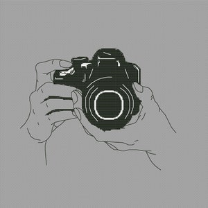 May include: A black and white line art illustration depicting a digital camera being held. The drawing features a detailed camera body with a prominent lens and shutter button, rendered in a clean, minimalist style against a solid grey background.