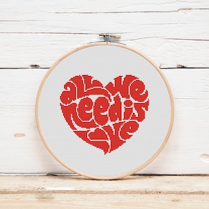 Könnte beinhalten: Ein roter, herzförmiger Stickrahmen mit den Worten "All we need is love", die in roter Farbe gestickt sind.