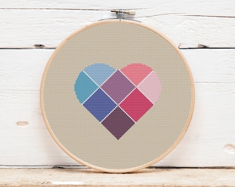 Puede incluir: Un bastidor de bordado de punto de cruz con un diseño de corazón. El corazón está compuesto por cuadrados en tonos azules, morados y rosas. El fondo es de tela beige y el bastidor es de madera. El diseño se muestra sobre una superficie de madera.